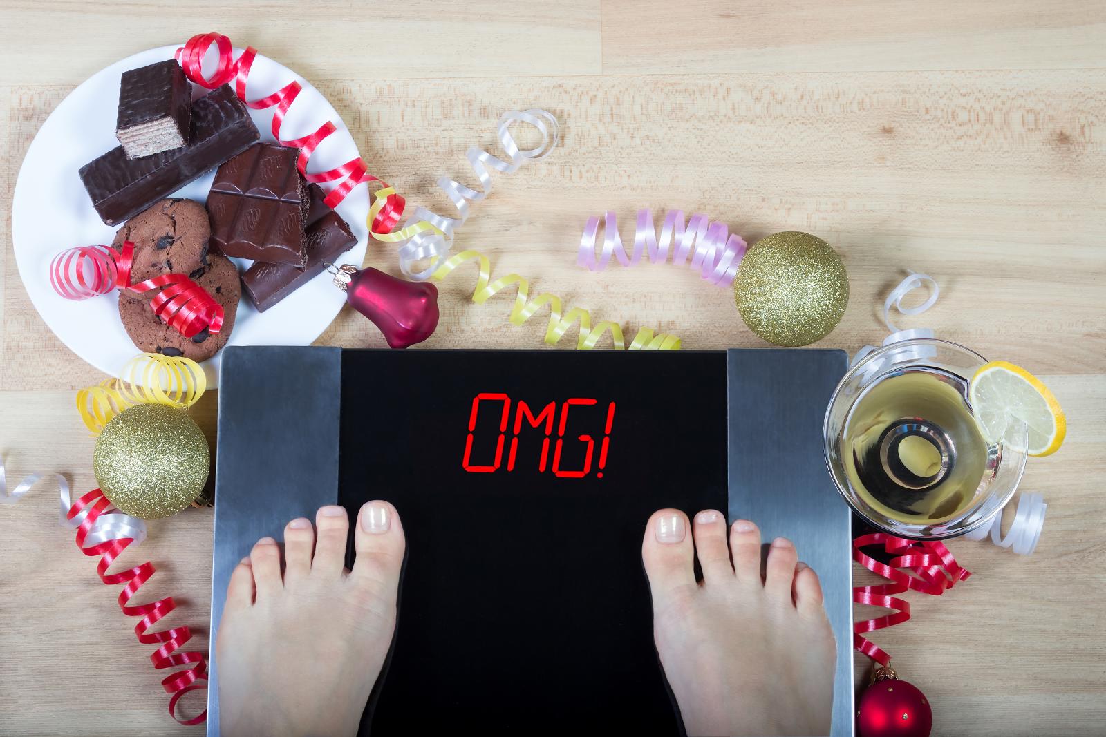 Digital scales with woman feet on them and sign"OMG!" surrounded by christmas decorations, sweets and alchohol. Demonstrates consequences of surfeit and eating unhealthy food during Christmas holidays.