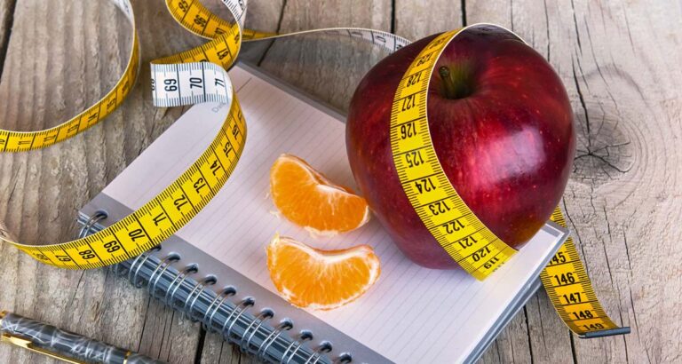 Apple wrapped in a measuring tape beside orange slices and a notebook